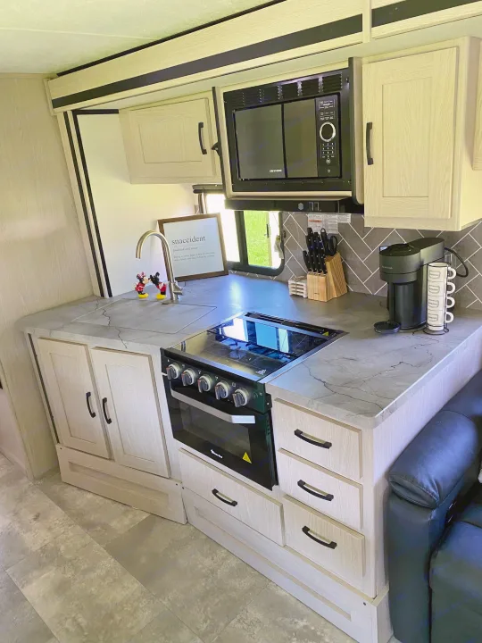 This modern RV rental kitchen features light wood cabinets, gray counters, microwave, stove, and sink, for comfortable travel.. Forest River two bedroom Apex ultralite 2022