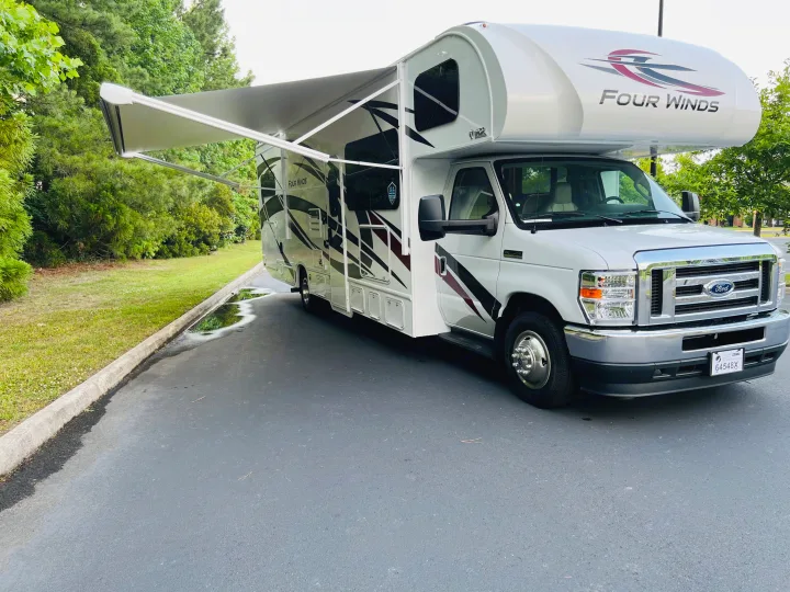 A white Four Winds RV with an extended awning is parked on asphalt with green trees and grass nearby. Ideal for rv rental.. Four Winds 31E 2022