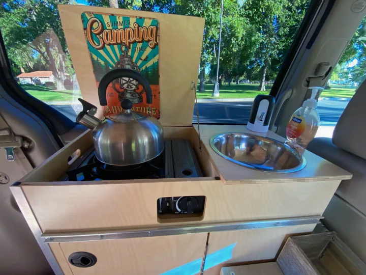 Inside an RV rental: a wooden camp kitchen setup with a sink, kettle on a stove, and soap dispensers, ready for camping trips.. Custom Camper 2004