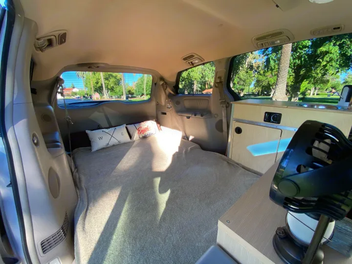 Inside an rv rental: a comfy bed, custom wooden kitchenette with sink, and fan, looking out to sunny park trees.. Custom Camper 2004