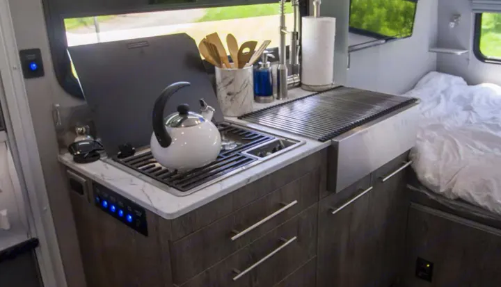 Modern RV rental kitchen with stove, sink, and kettle; bed nearby for compact living.. Intech Sol Horizon Rover 2022