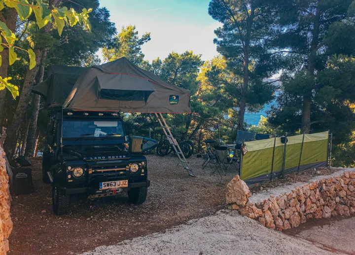 Overland camping setup: a black Land Rover Defender RV rental with a roof tent, parked by the sea amidst pine trees.. Land Rover Defender 110 XS Utility + 2 Roof Tents 2013