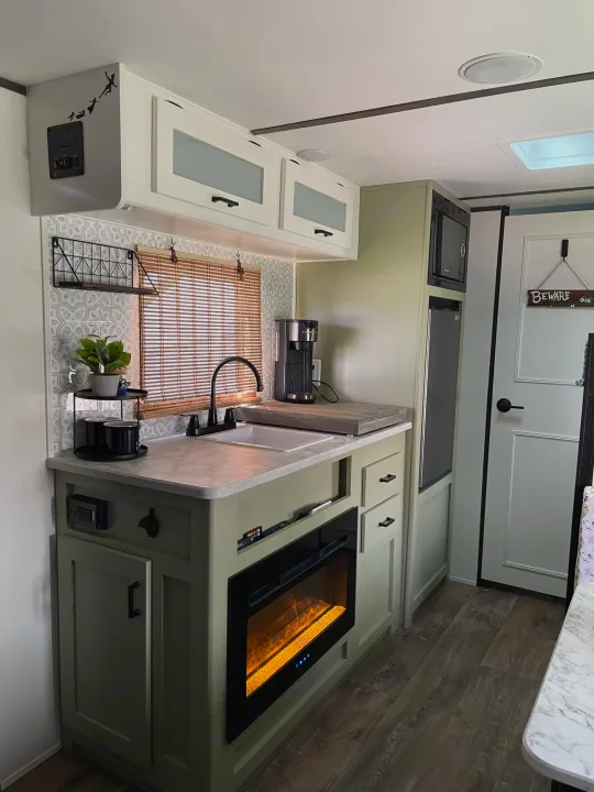 Modern RV rental kitchen with sage cabinets, white counters, and electric fireplace, perfect for compact living.. Dutchmen Coleman Lantern LT 2021