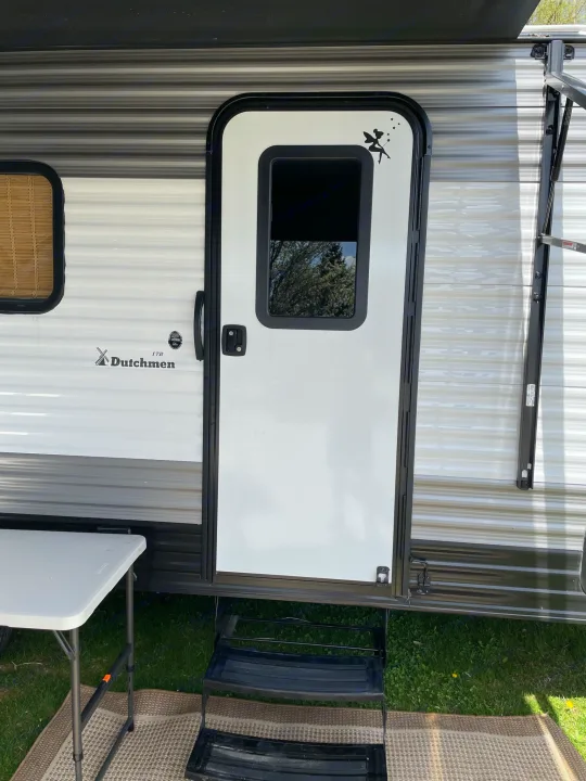 Experience an RV rental! This image shows the RV entrance with steps, a folding table, and outdoor mat, ready for camping.. Dutchmen Coleman Lantern LT 2021