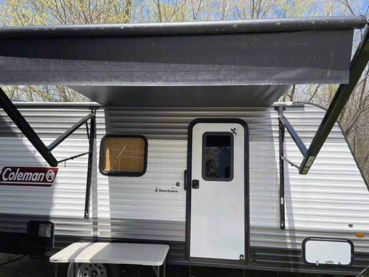 Close-up of an RV rental: a white Coleman Dutchmen travel trailer with an extended awning at a sunny campsite.. Dutchmen Coleman Lantern LT 2021