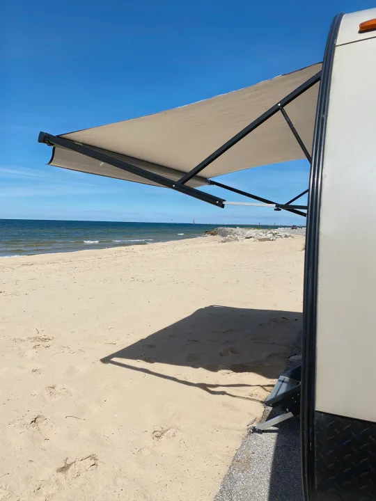 An RV rental with its awning extended, casting a shadow on a sunny beach next to the blue sea. A road and pier are visible in the distance.. Forest River Wildwood X-Lite 2019