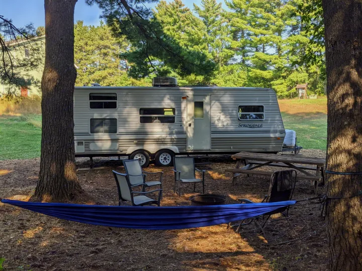 This RV rental campsite features a Springdale travel trailer, blue hammock, fire pit, chairs, and picnic table in a wooded setting.. Keystone RV Springdale 2003