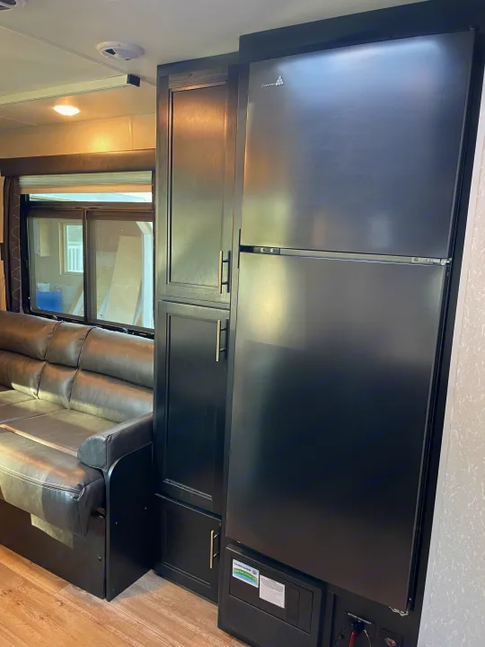 Inside a dark-toned RV rental: features a large black refrigerator, sleek cabinets, and a comfortable leather sofa next to a window.. Forest River Cherokee Grey Wolf 2021