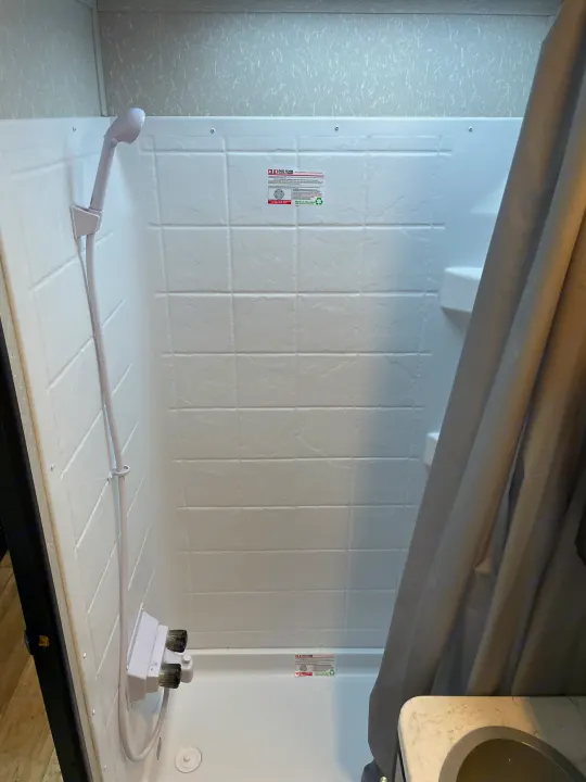 Interior of a white RV rental shower with a handheld shower and control knobs. Includes a small shelf.. Forest River Cherokee Grey Wolf 2021
