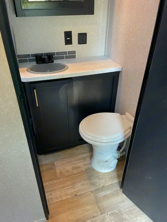 Compact RV rental bathroom showcasing a modern dark vanity, sink, white toilet, light wood flooring, and gray walls.. Forest River Cherokee Grey Wolf 2021
