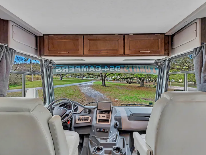 Spacious RV rental interior showing the driver's area, dashboard, and a clear view of a park through the windshield.. Coachmen Mirada 2021