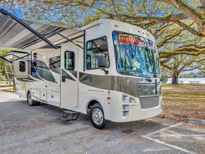 A cream-colored Mirada RV rental with an extended awning is parked on pavement near a lake and large mossy trees.. Coachmen Mirada 2021