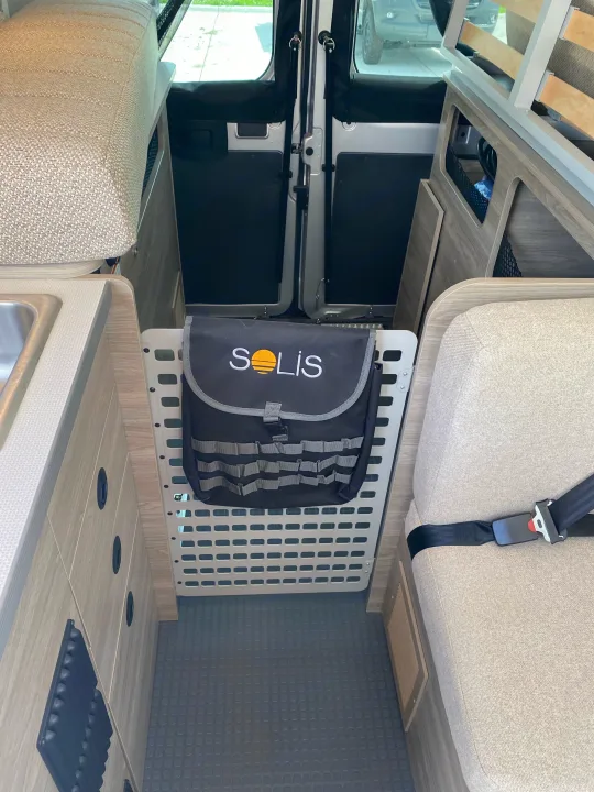RV rental interior: kitchen sink, cushioned seating with seatbelt, and a "SOLIS" storage bag on a grid panel inside a camper van.. Winnebago pocket 2023