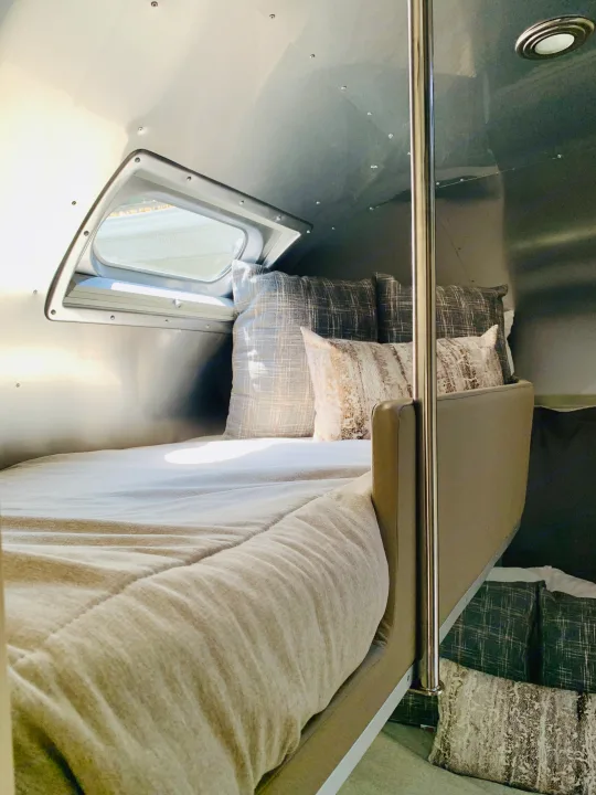 Inviting RV rental interior: a plush bed with patterned pillows, bright window, and sleek metallic walls. A second bunk is visible.. Airstream Flying Cloud 2022