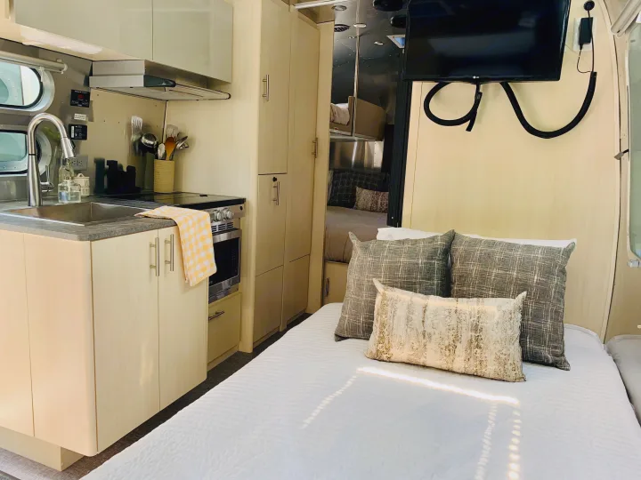 Modern RV rental interior: a compact kitchen with sink/stove, ample cabinets, and comfortable bed with pillows & TV.. Airstream Flying Cloud 2022