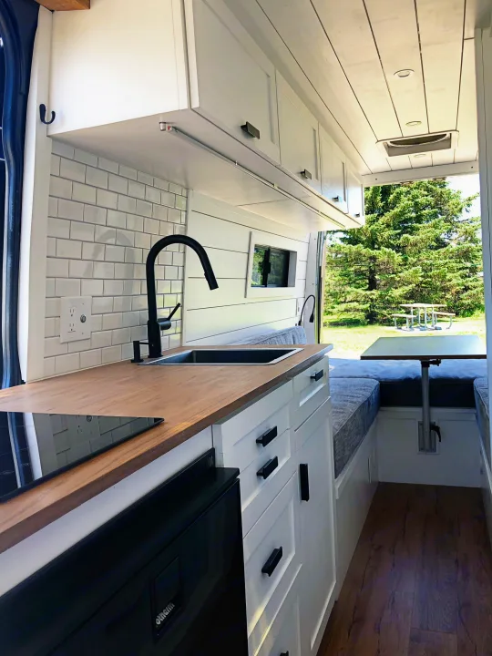 Bright RV rental interior: modern kitchen with wood counters, subway tile, sink, and a convertible dining/lounge area for mobile living.. Mercedes-Benz Sprinter 2015
