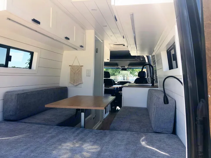 View inside a bright, modern RV rental camper van. Features a dining area with gray benches, kitchen, & overhead cabinets.. Mercedes-Benz Sprinter 2015