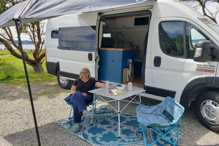 A woman unwinds beside her white rv rental, a converted Ram ProMaster van, with an outdoor snack setup by the water.. Dodge Promaster 2500 2021