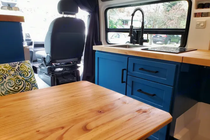 Modern **rv rental** kitchen & dining in a custom van conversion, showcasing blue cabinetry, wood counters, and swiveling seat.. Dodge Promaster 2500 2021