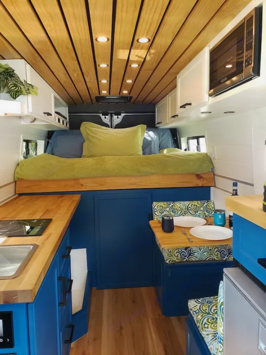 Inside a well-appointed RV rental van: custom blue cabinetry, wood ceiling, kitchen, bed, and dining area.. Dodge Promaster 2500 2021