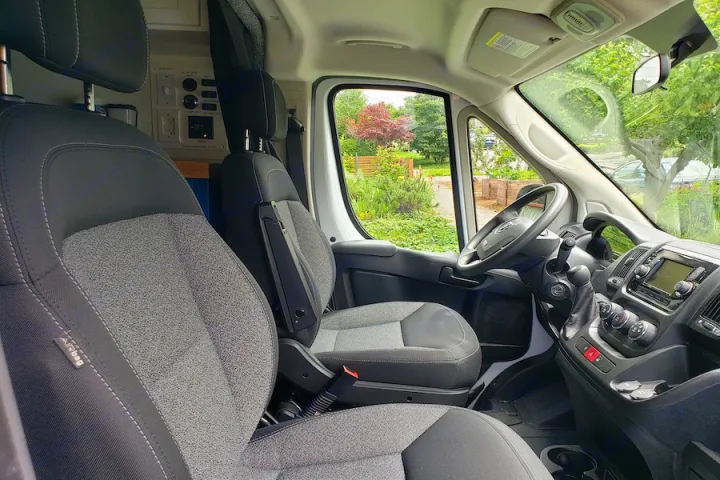 Inside an RV rental: spacious front cabin with gray driver/passenger seats, dashboard, and controls. Green garden visible through windows.. Dodge Promaster 2500 2021