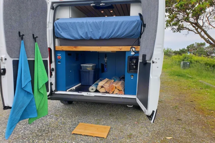 Back of RV rental van open: upper bed, lower blue storage with firewood & bins, towels on doors, & ground mat. Camping ready!. Dodge Promaster 2500 2021