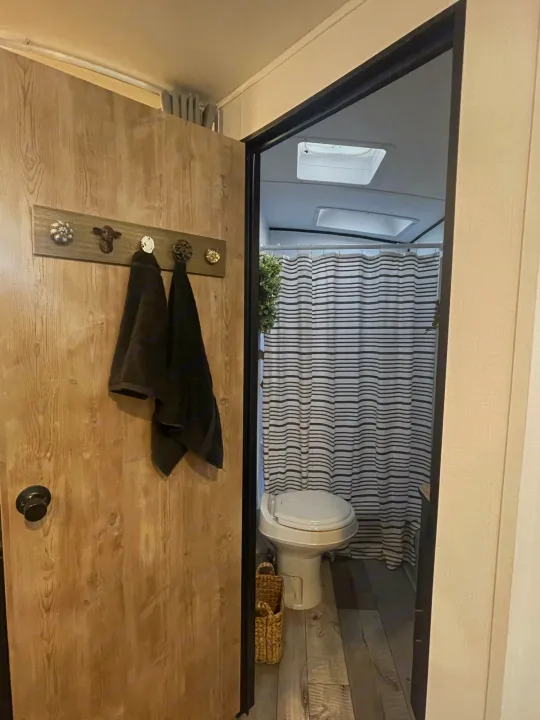 RV rental bathroom interior: toilet, striped shower curtain, and wood-look floor. Towels hang on door's decorative rack.. Keystone Springdale 2022
