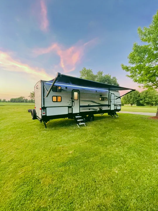 This RV rental travel trailer is ready for adventure, set in a lush grassy field under a vibrant pink sunset sky with its awning lights on.. Keystone Springdale 2022