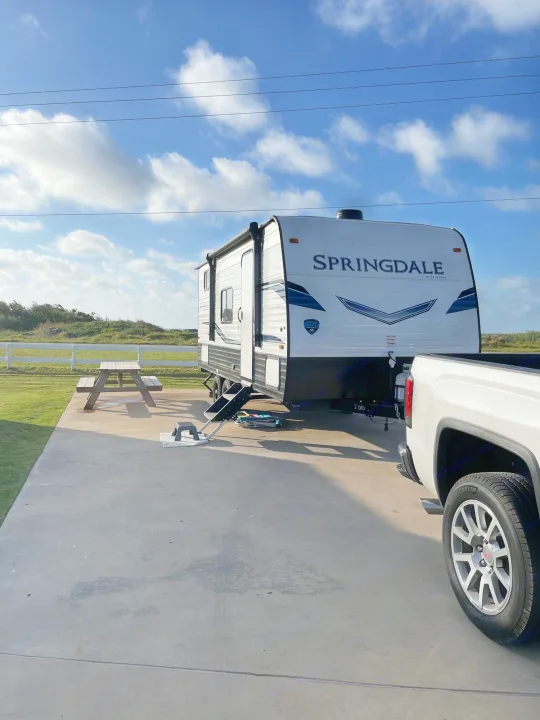 An RV rental is parked on a concrete pad with a picnic table and white truck, under a blue sky at a grassy campsite.. Keystone Springdale 2022