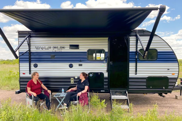 Two women relax outside a blue and white Wolf Pup RV rental with an extended awning, enjoying an outdoor camping trip.. Forest River Cherokee Wolf Pup 2022