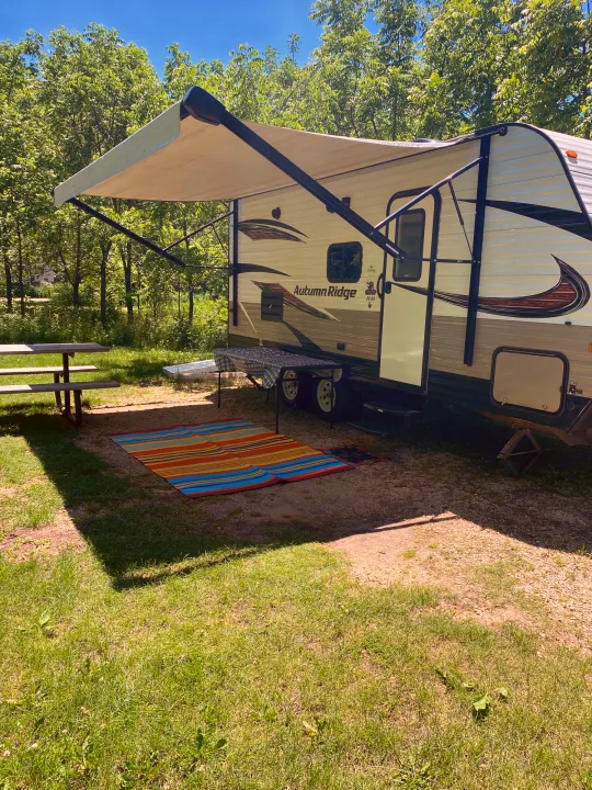 Perfect RV rental outdoor setup: travel trailer with awning, picnic table, and colorful mat at a quiet campsite.. Starcraft Autumn Ridge 2018