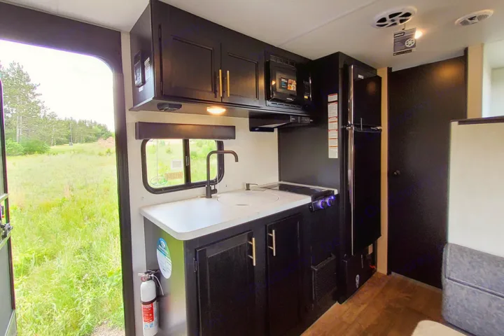 RV rental interior: A modern, compact kitchen with black cabinets, white counter, sink, microwave, and fridge. Open door view of grassy outdoors.. Forest River Cherokee Wolf Pup 2022