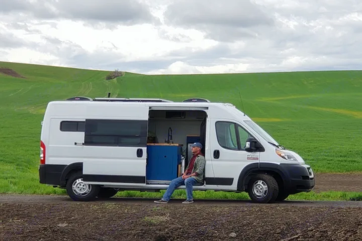 A white RV rental van with an open side door, showcasing a man sitting inside amidst green hills.. Dodge Promaster 2500 2021