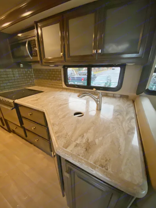 Modern RV rental kitchen: Stone countertop, sink, dark wood cabinets, subway tile backsplash, and window view.. Flair-Fleetwood Flair-Fleetwood Motorhome 2020