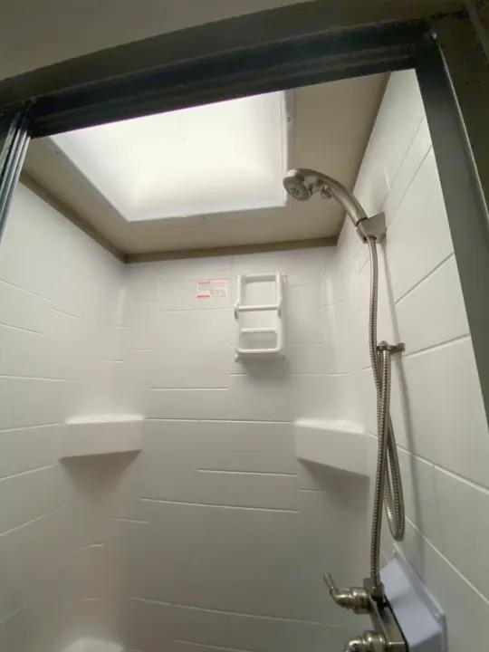 View into a compact RV rental shower featuring a handheld showerhead, white tiled walls, and a bright overhead skylight.. Flair-Fleetwood Flair-Fleetwood Motorhome 2020