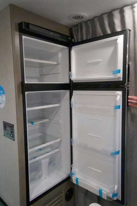 Two-door RV rental fridge and freezer, open to show clean, empty shelves and door bins, still with blue securing tape.. Coachmen Freelander 2021