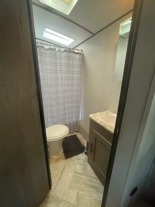 Interior view of a compact RV rental bathroom, featuring a toilet, shower with a striped curtain, and vanity with a sink.. Keystone RV Springdale 2021