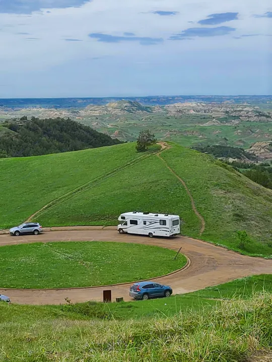 Scenic overlook with an RV rental on a winding dirt road. Green hills, distant badlands, and two cars complete the natural view.. Forest River Sunseeker 2017