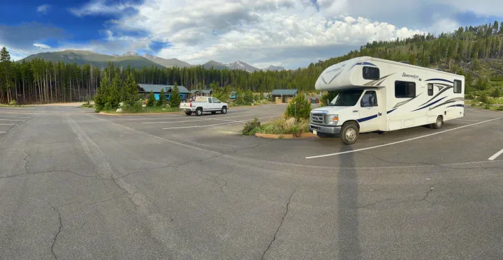 White Sunseeker RV rental and a pickup truck parked in a large mountain forest lot, with peaks and buildings in the background.. Forest River Sunseeker 2017