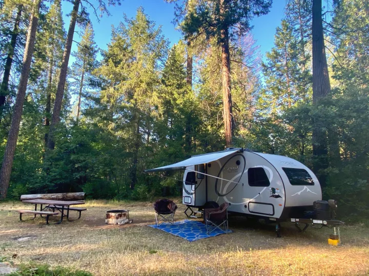A cozy R-Pod rv rental set up at a forest campsite with chairs, a mat, and a fire pit, ideal for outdoor living.. R-Pod R-Pod Trailer 2021