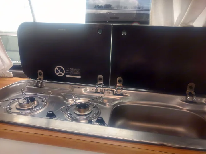 Inside an RV rental: a Dometic compact kitchen with a two-burner gas stove and sink, featuring partially open glass covers.. Volkswagen Westfalia 1986