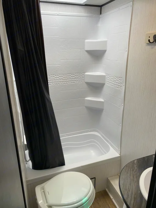 RV rental bathroom features a clean white shower/tub with shelves, a black curtain, and toilet.. Heartland RVs Trailrunner 2021