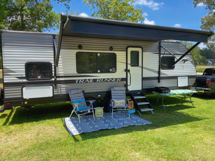 Relaxing RV rental setup: a parked travel trailer with extended awning, chairs, and table on a sunny green lawn.. Heartland RVs Trailrunner 2021
