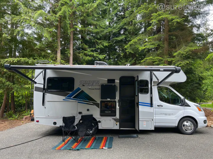 A white Cross Trail rv rental motorhome is set up at a treed campsite with awning extended, chairs, and mat.. Forest River Coachmen Cross Trail XL 2022