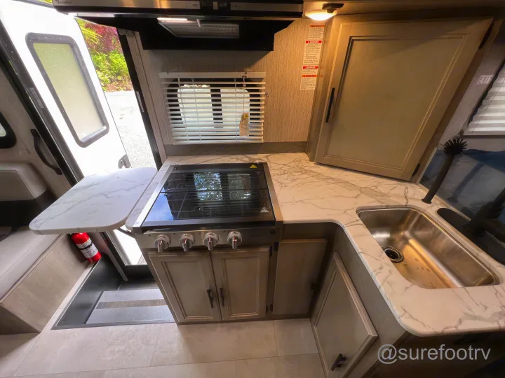 Compact RV kitchen: light wood cabinets, marble-look counter, stainless sink, and stove. All set for your next rv rental trip!. Forest River Coachmen Cross Trail XL 2022