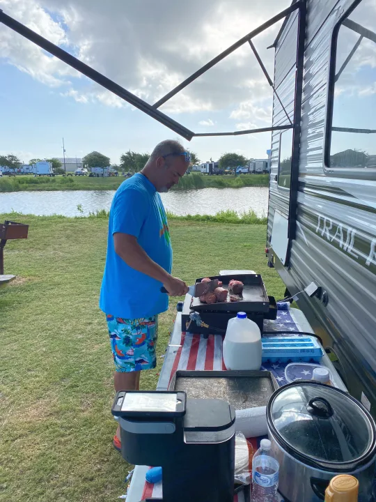 Man cooking on a griddle outside his RV rental at a lakeside campsite, with food prepped on an American flag table.. Heartland RVs Trailrunner 2021
