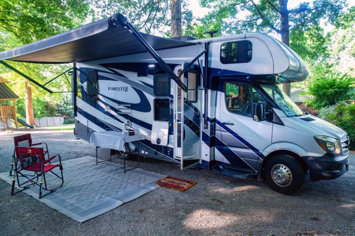 RV rental set up at a shaded campsite with awning, outdoor rug, table, and chairs.. Forest River Forester MBS 2018