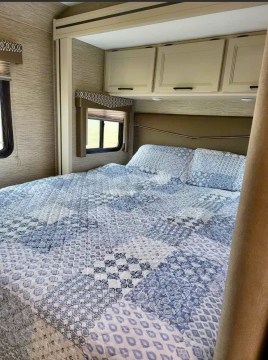 Cozy RV rental bedroom featuring a bed with patterned blue and white bedding, overhead cabinets, and windows.. Thor Freedom Elite 2022