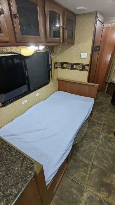 RV rental interior. This bedroom has a bed with light blue sheet, wooden cabinets above, a window, and tiled floor.. Heartland RVs North Trail 2013