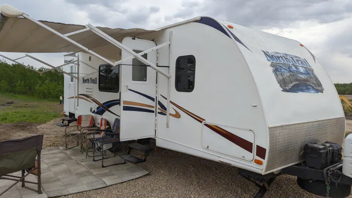 An RV rental with an extended awning, chairs, and mat at a gravel campsite. Ready for outdoor adventure and travel.. Heartland RVs North Trail 2013